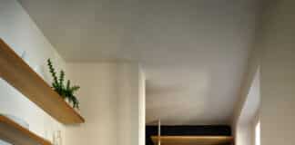 A modern kitchen with wood cabinets, open shelves, plants, and a terrazzo floor. A round white table with a curved bench holds a bowl of oranges, a vase, and decor. Shelves in a dark alcove display books and plants.