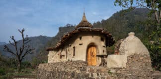 A small, rustic house with curved walls, a thatched conical roof, and a wooden door sits atop a stone wall with steps, surrounded by greenery and mountains in the background.