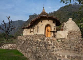 A small, rustic house with curved walls, a thatched conical roof, and a wooden door sits atop a stone wall with steps, surrounded by greenery and mountains in the background.