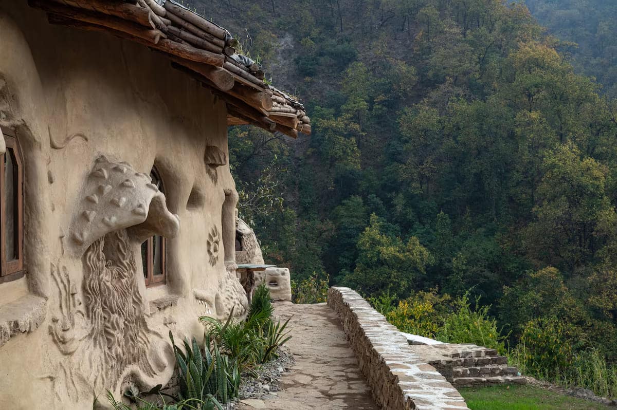 A rustic house with walls featuring sculpted mushroom designs stands beside a stone path and wall, overlooking a forested hillside with dense green trees.