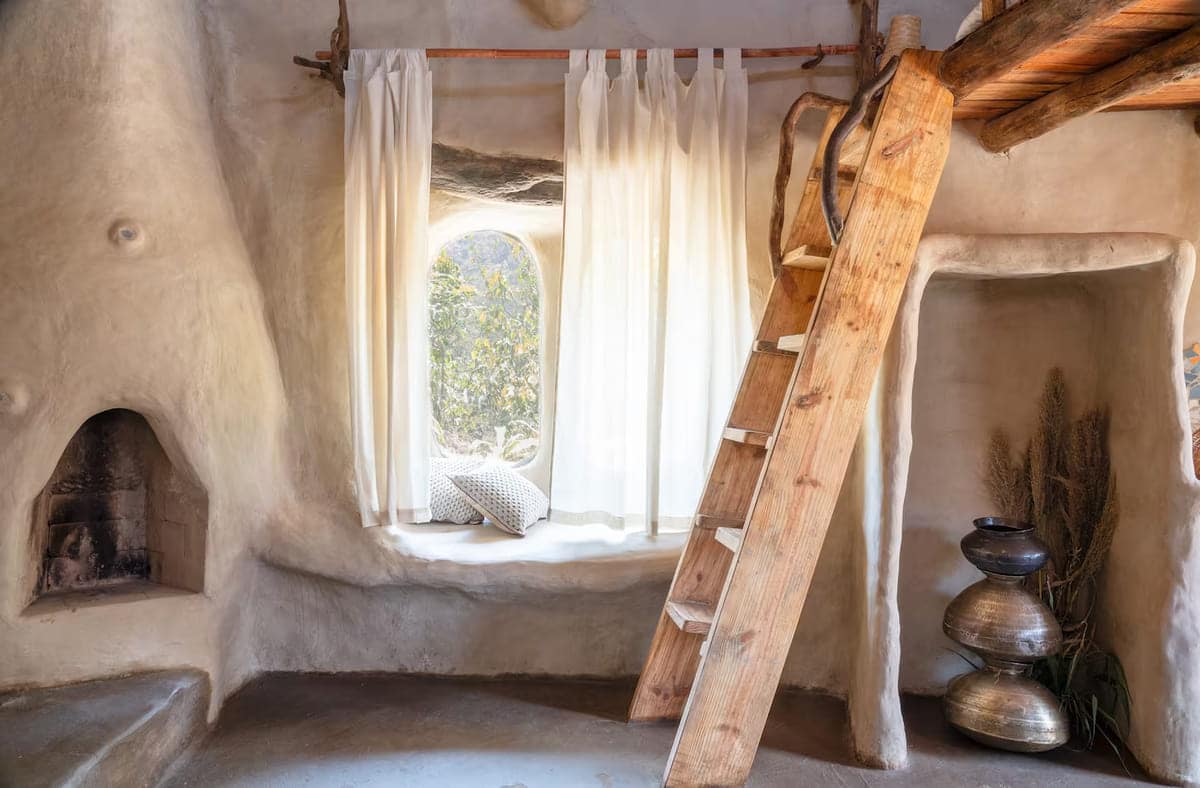 Cozy, rustic room with curved, earth-toned walls, a built-in fireplace, a wooden ladder leading to a loft, a window with sheer curtains, a cushioned window seat, and metallic vases on the floor.