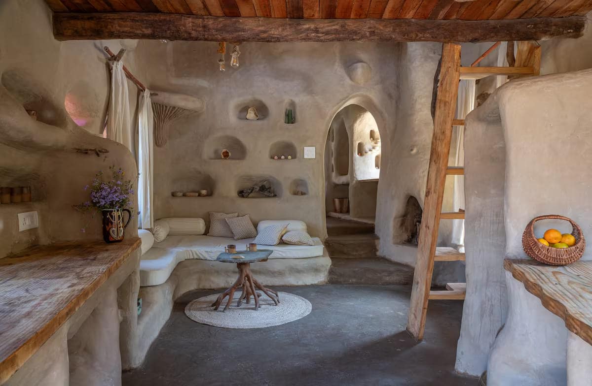 A cozy, rustic room with curved earthen walls, built-in shelves, a small sitting area with cushions, wooden ceiling beams, a ladder leading to a loft, and natural light streaming through curtained windows.