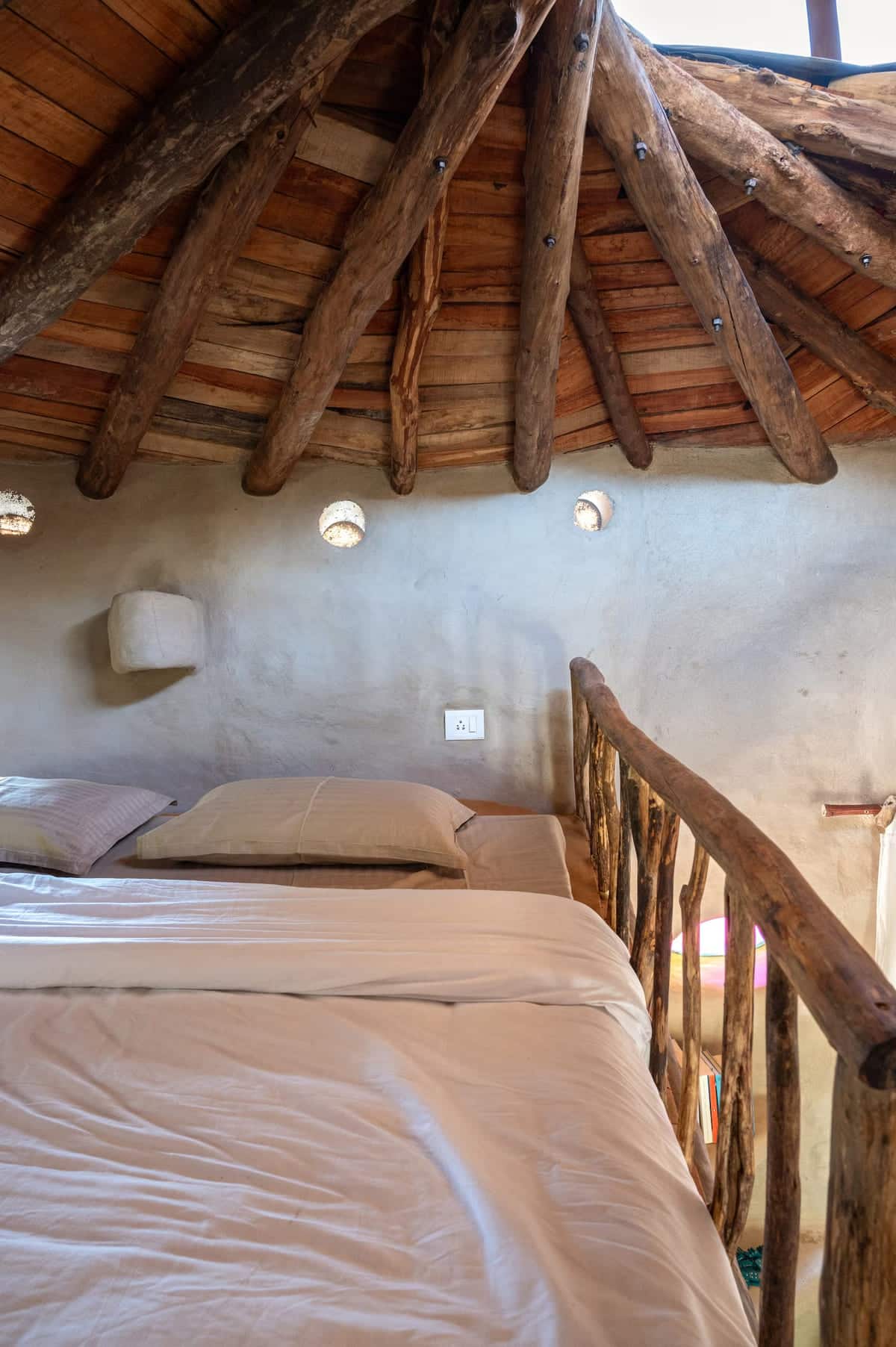 A cozy loft bed with beige bedding sits under a rustic wooden ceiling with exposed beams. The space features small round windows and a natural wooden railing, creating a warm, earthy atmosphere.