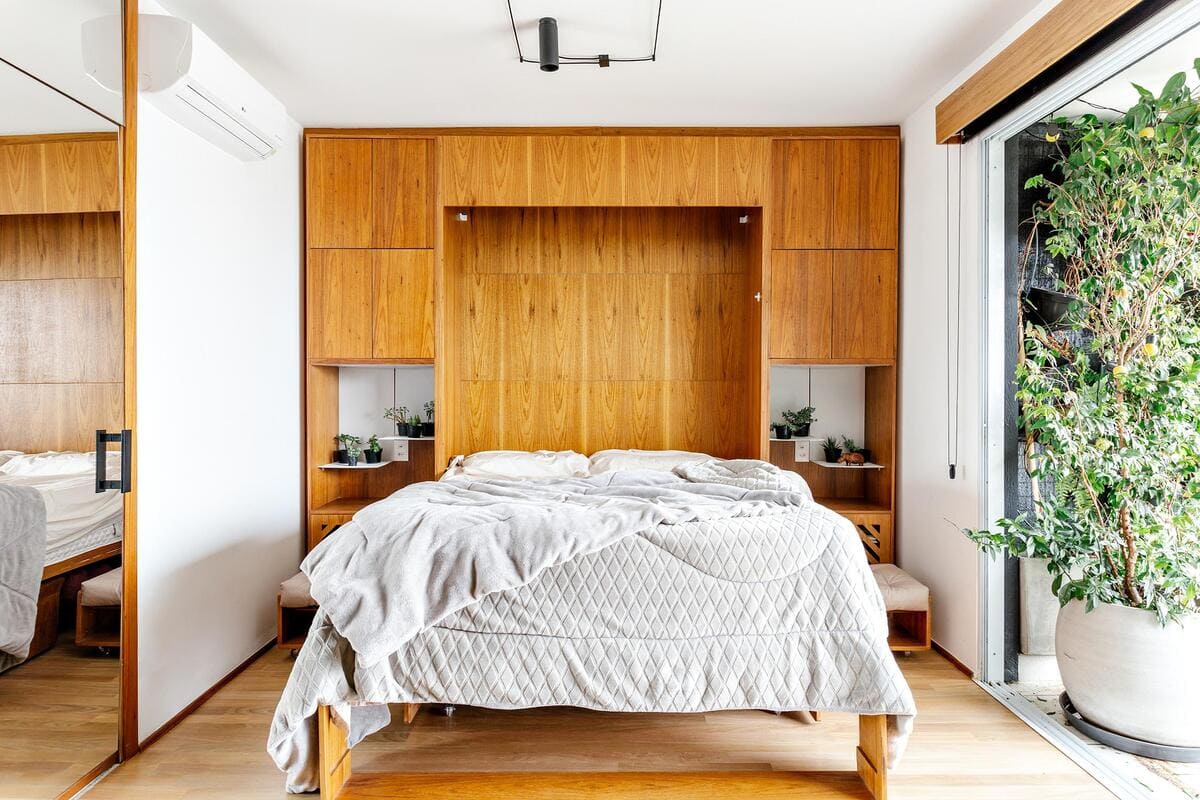 A modern bedroom with a wooden headboard and built-in shelves, an unmade bed with light bedding, and a large potted plant near a sliding glass door bringing in natural light.