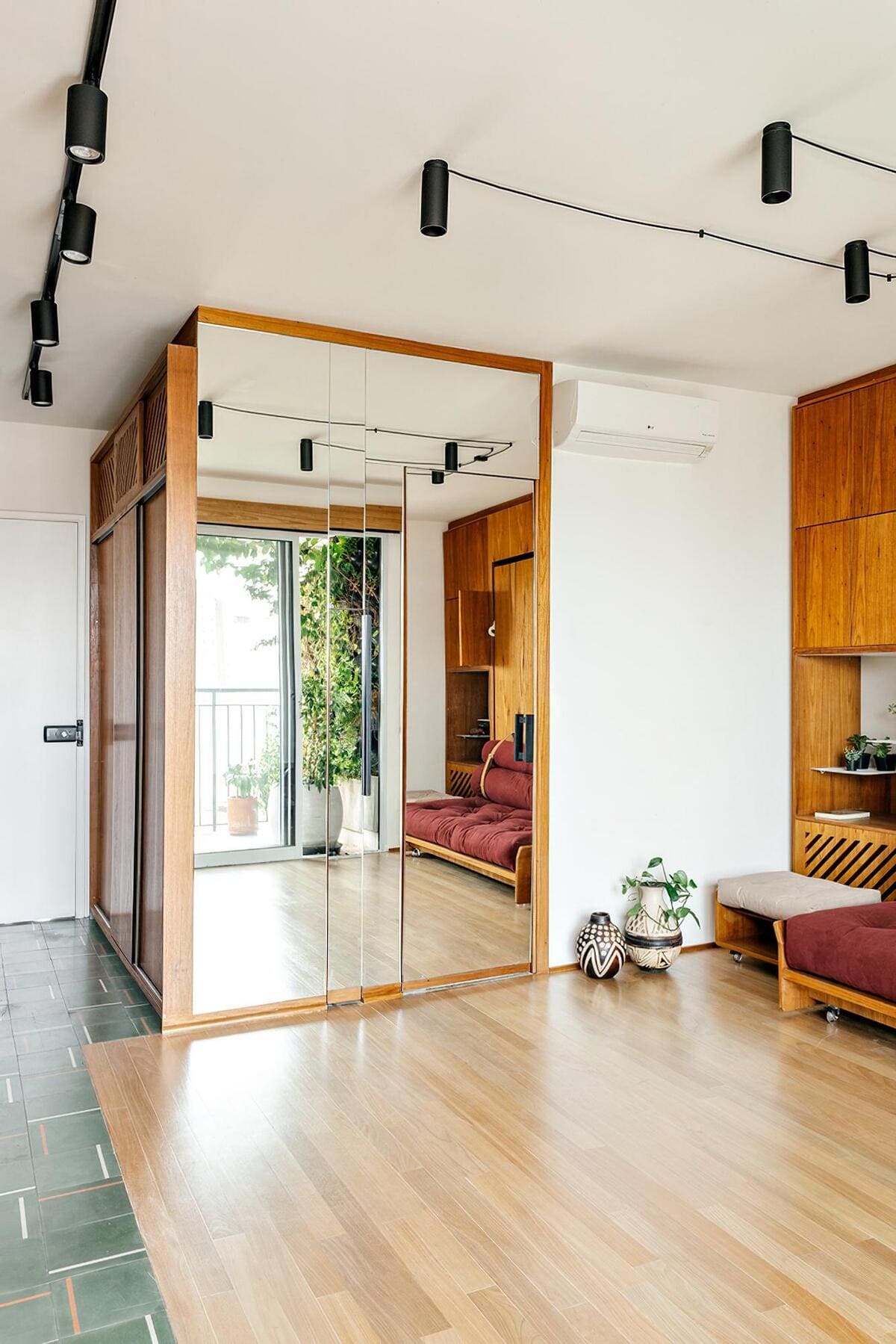 A modern living room with light wood flooring, mirrored closet doors, track lighting, a maroon sofa, plants, and a balcony visible through a glass door. The decor is minimalist with wood accents.