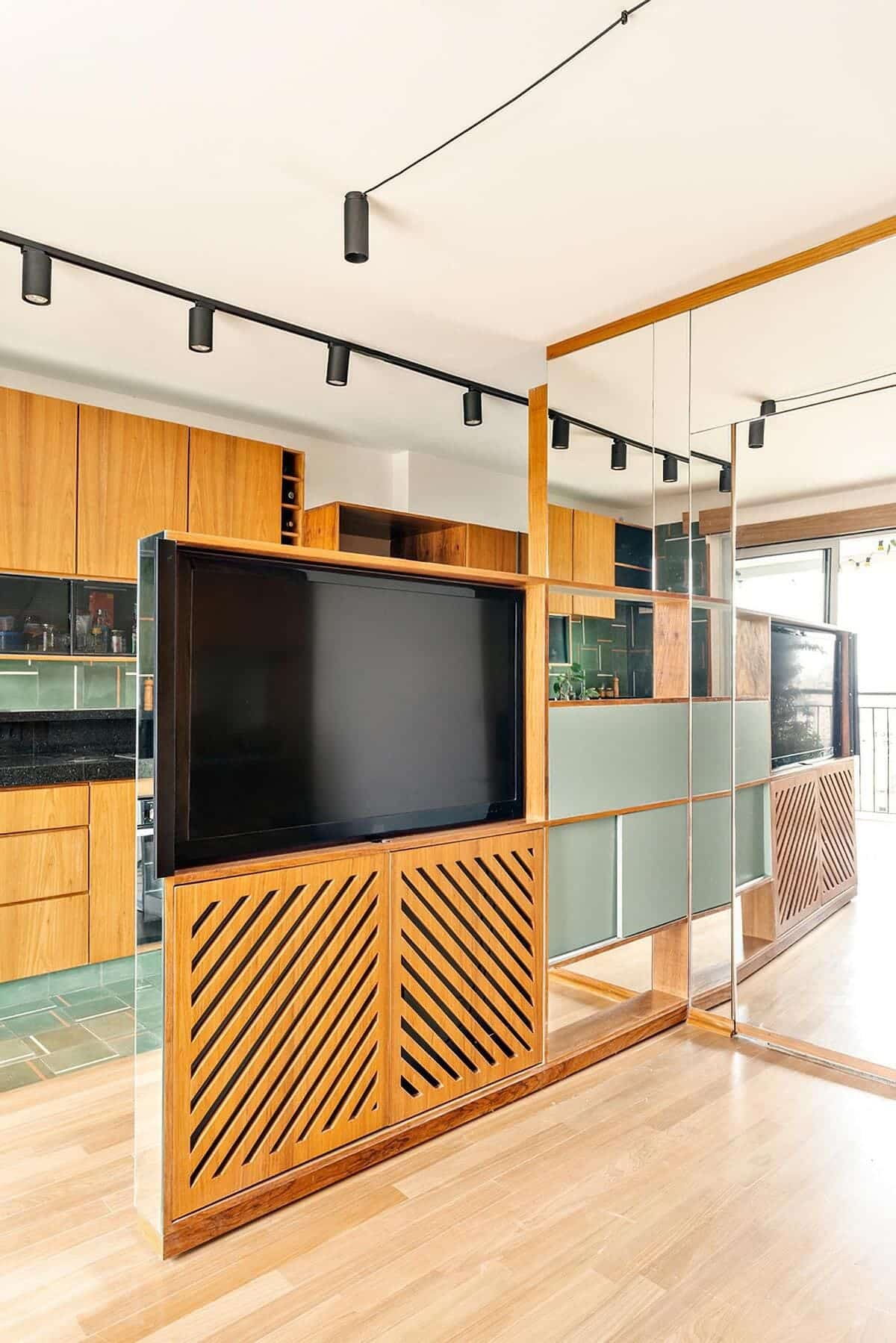 A modern living space with wood flooring, a large flat-screen TV in a wooden entertainment unit, mirrored wall panels, and a kitchen with wood cabinets and green tiles in the background.
