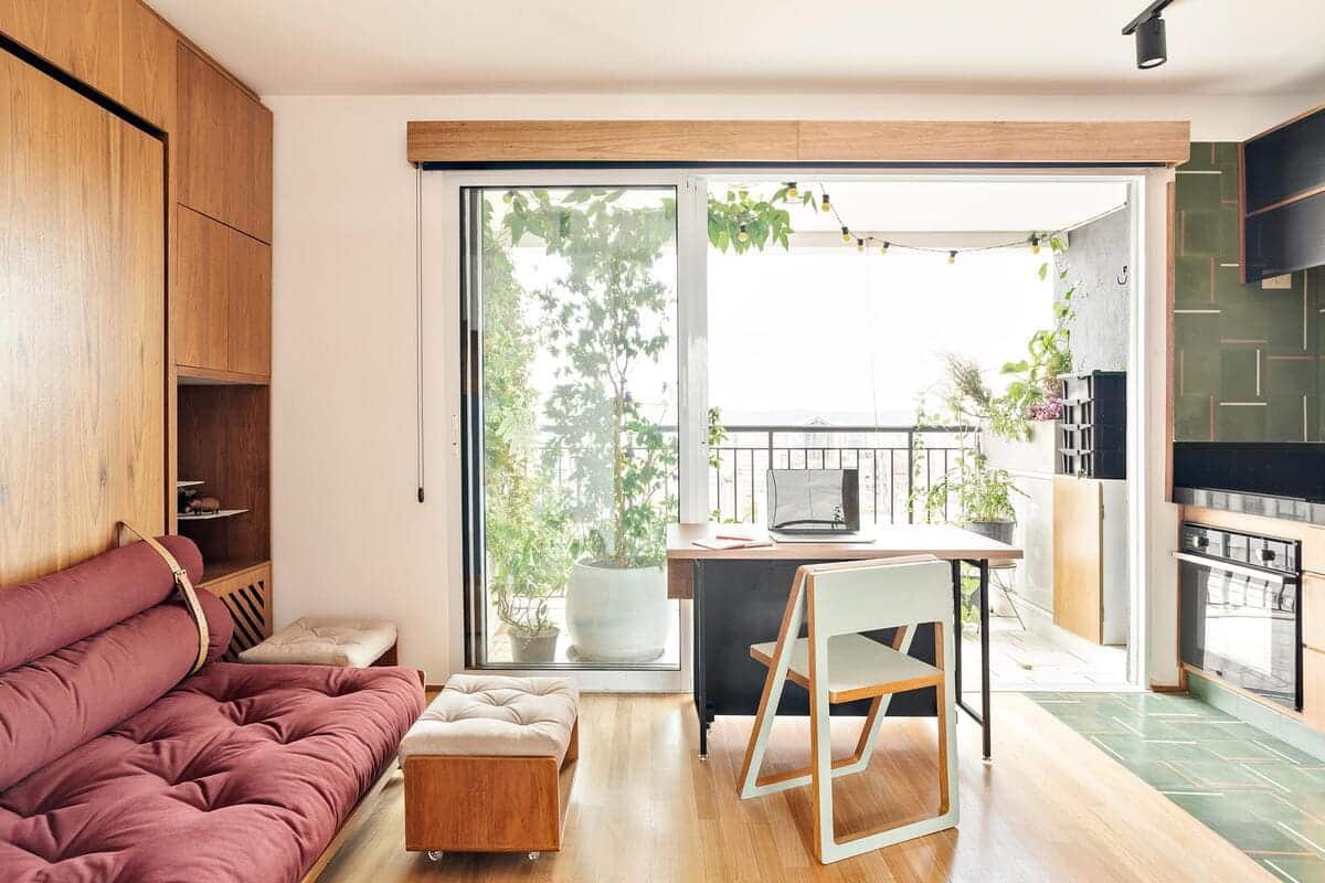 A bright, modern living room with a maroon sofa, wooden furniture, a small desk and chair by large glass sliding doors, and a balcony with plants visible outside. Natural light fills the space.
