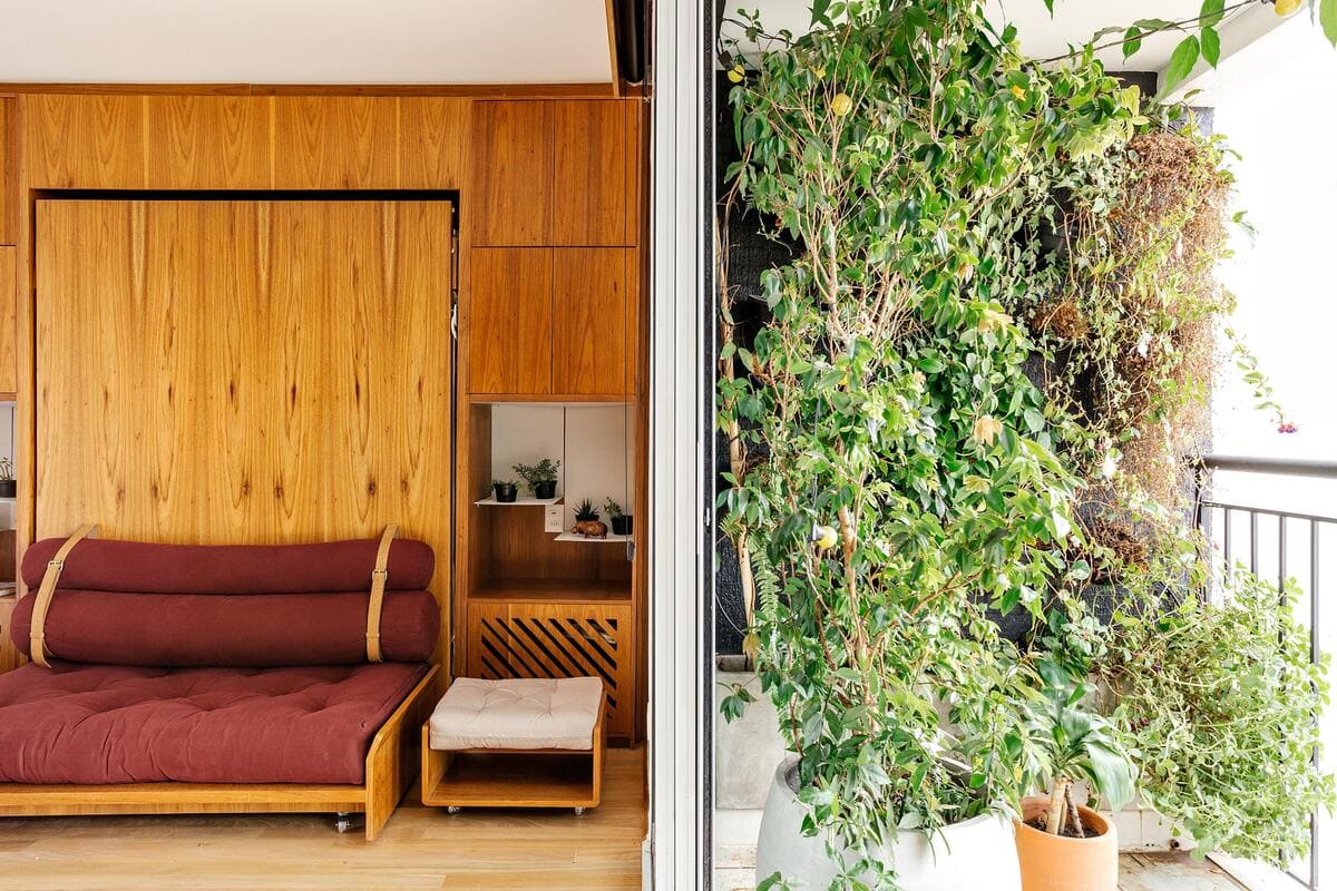 A cozy room with a wooden wall and futon sofa sits next to a balcony filled with green potted plants, separated by a sliding glass door.