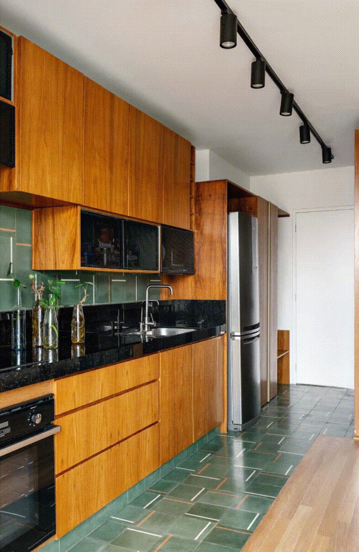 Modern kitchen with wooden cabinets, black countertops, green tiled backsplash, stainless steel refrigerator, oven, and sink. The floor features green tiles with a wooden strip, and black track lighting is on the ceiling.