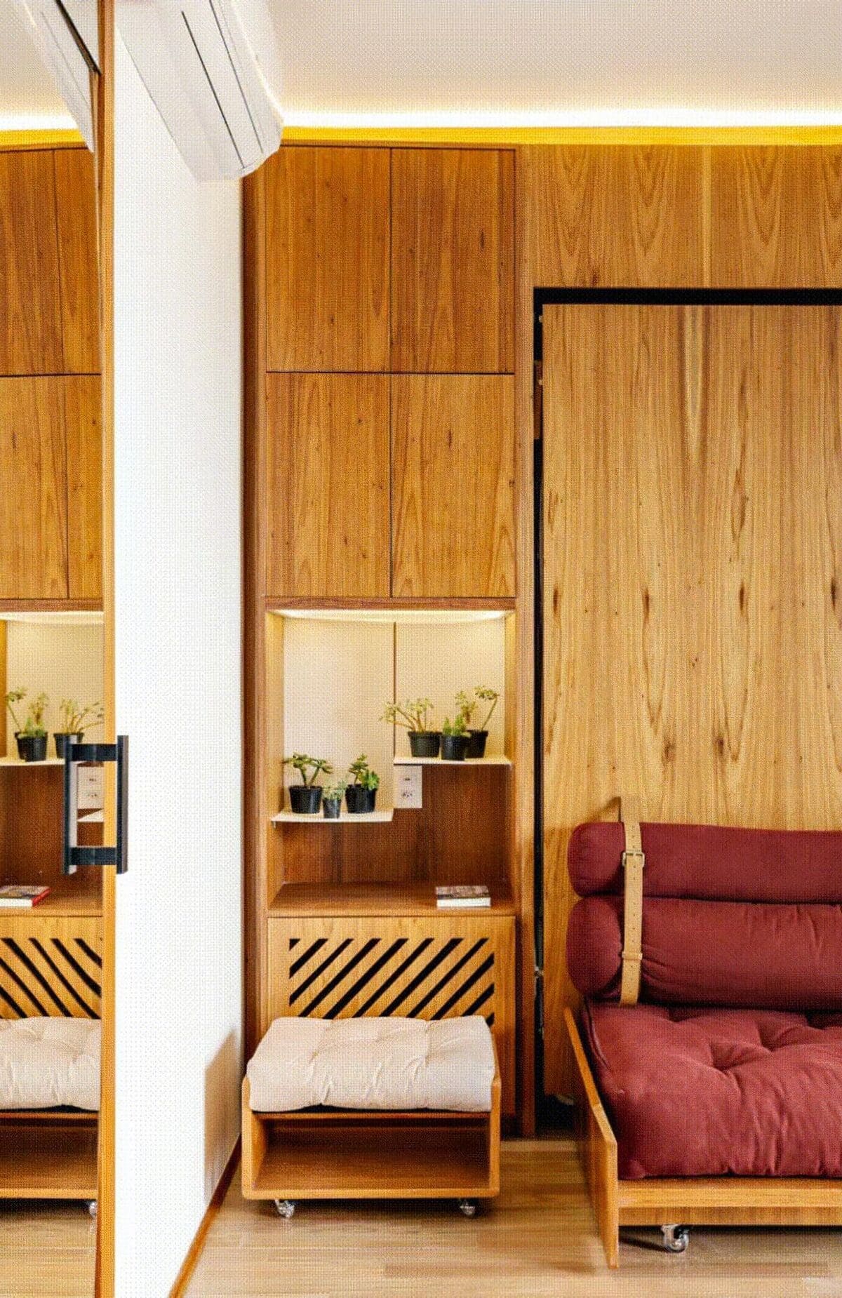 A modern room with wooden walls and furniture, featuring a maroon cushion sofa, a small stool, potted plants on a shelf, and a large mirror reflecting part of the interior. Warm lighting enhances the cozy atmosphere.