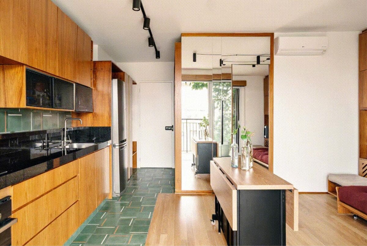 Modern apartment with wooden cabinets, green tiled kitchen floor, a sleek black countertop, and minimalist decor. A large mirror and sliding door open to a balcony with natural light and plants.
