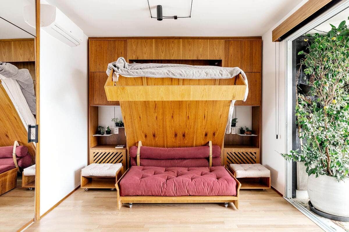 A modern room with a wooden Murphy bed partially folded down over a red sofa, shelves with plants on each side, and a large potted plant by a sliding glass door letting in natural light.