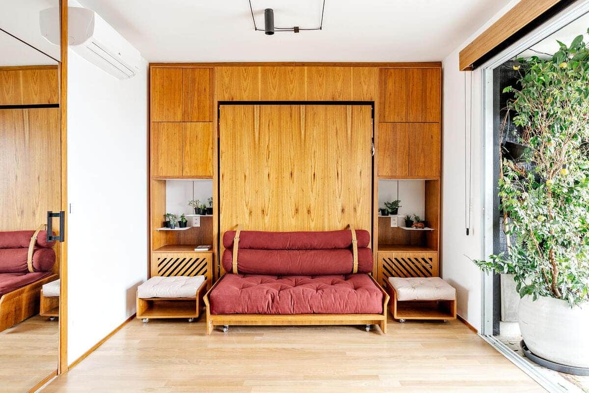A modern living room with a wooden wall unit, a red cushioned sofa, two small side tables with potted plants, light wood flooring, and a large potted plant beside a sliding glass door.