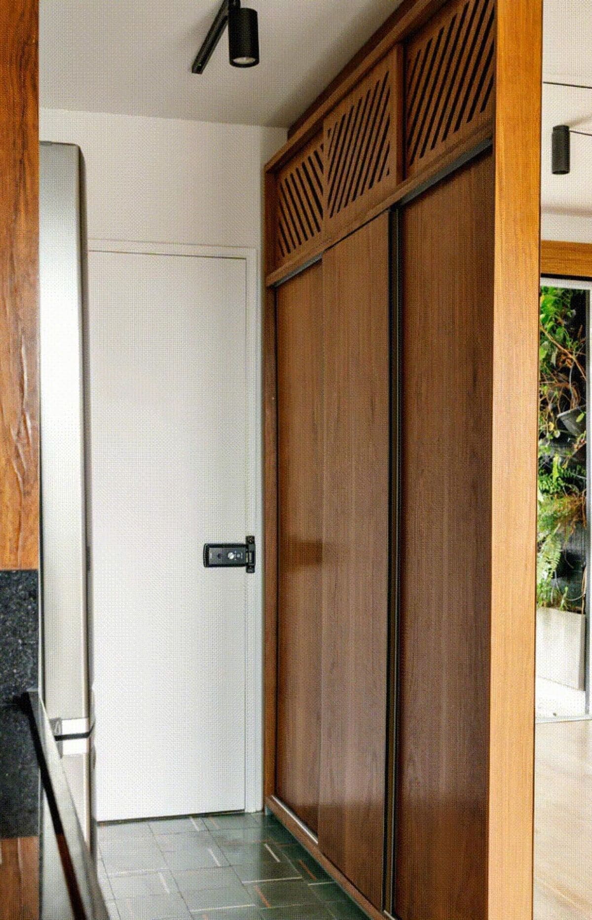 A hallway with green tile flooring, a white door with a black handle, wooden sliding closet doors on the right, and a partial view of a kitchen counter on the left. A window reveals greenery outside.