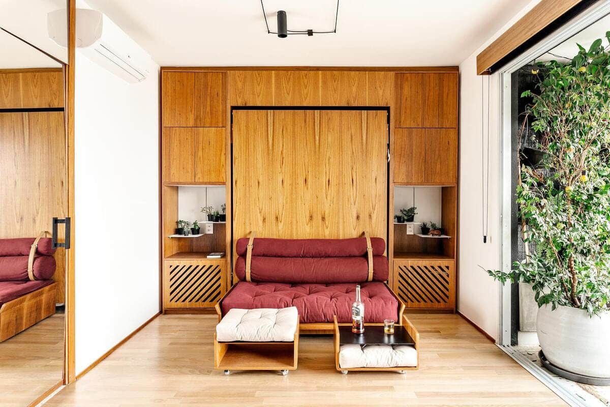 A modern living room with a wooden wall unit, red cushioned sofa, matching ottoman, small wooden coffee table, plants, and large sliding glass doors letting in natural light.