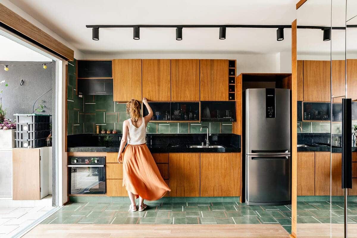 A woman in a white top and orange skirt stands in a modern kitchen with wood cabinets, green tile backsplash, a stainless steel fridge, and large windows letting in natural light.
