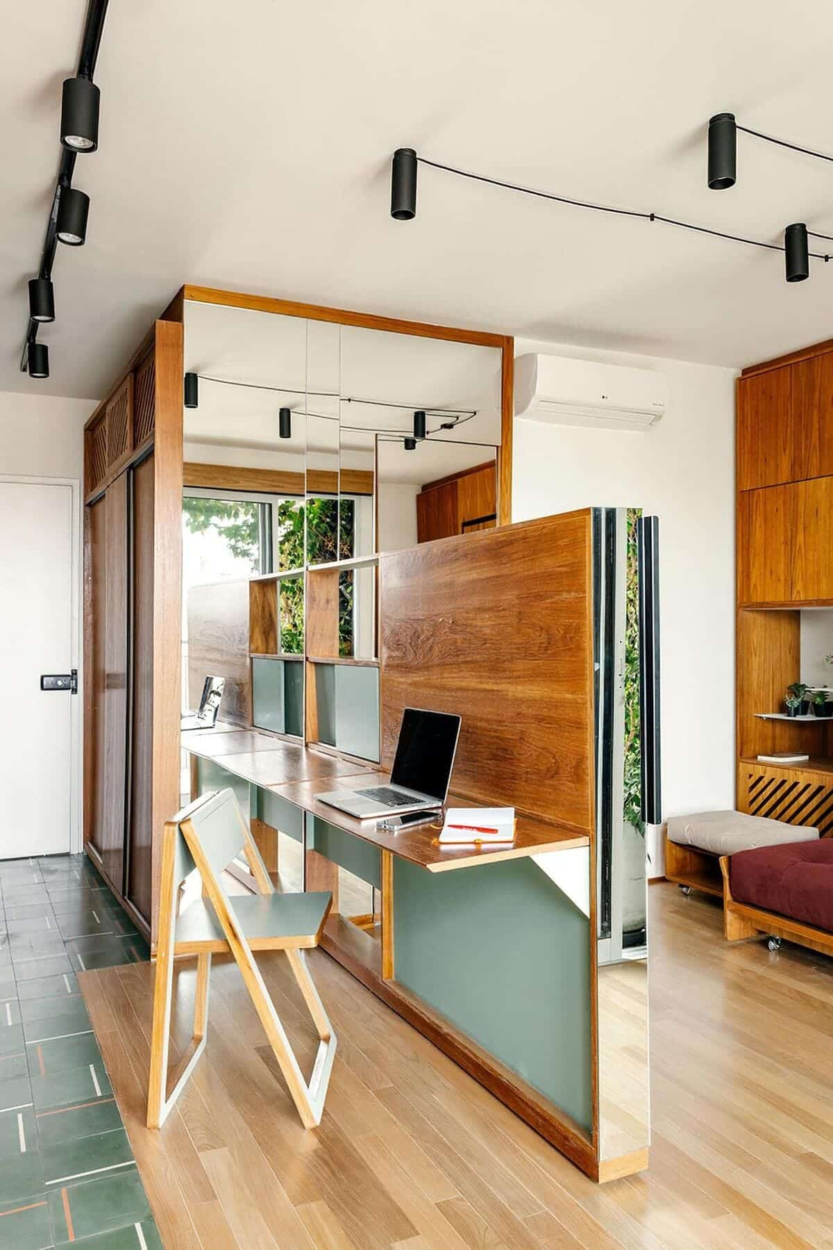 Modern home office with wood and green accents, featuring a long shared desk with three laptops, geometric chair, track lighting, mirrors, and large window letting in natural light.