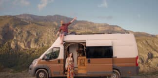 A woman stands by a brown and white camper van with a dog inside, while a man sits on the roof waving, set against a backdrop of mountains and a clear sky.
