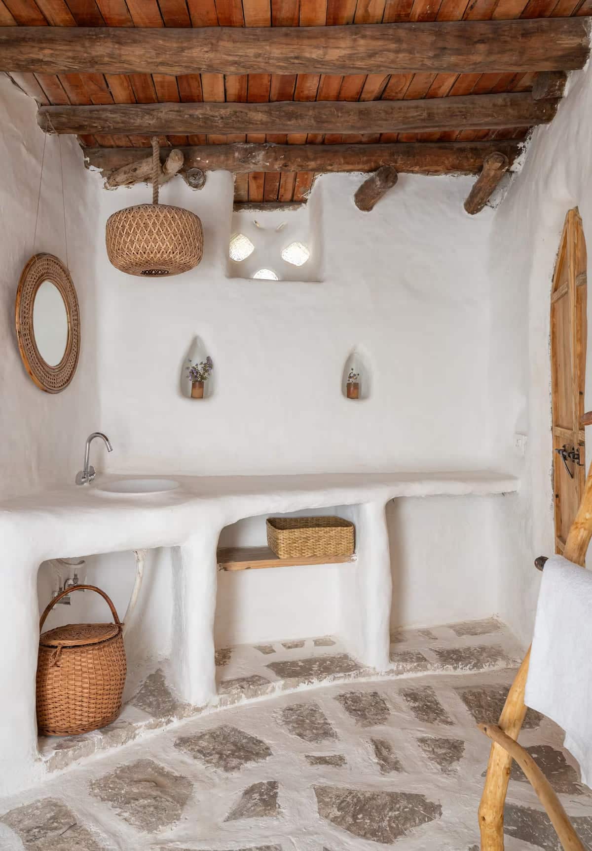 A rustic bathroom with white plastered walls, stone floor, wooden ceiling beams, a wicker pendant light, round mirror, built-in counter with a sink, and wicker baskets used for storage.