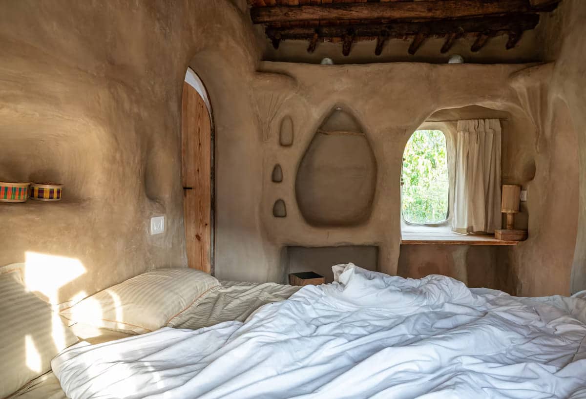 A cozy bedroom with an unmade bed, beige clay walls, a wooden door, built-in shelves, and a window with a curtain letting in natural light from outside greenery.