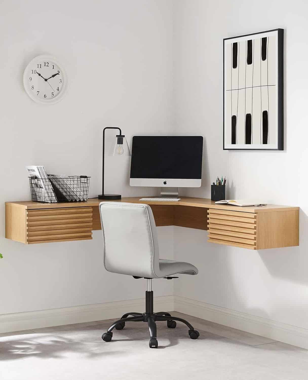A modern corner desk setup with a computer, desk lamp, and office chair. The floating wooden desk holds office supplies and a wire basket. A wall clock and a black-and-white art piece hang on the white walls.