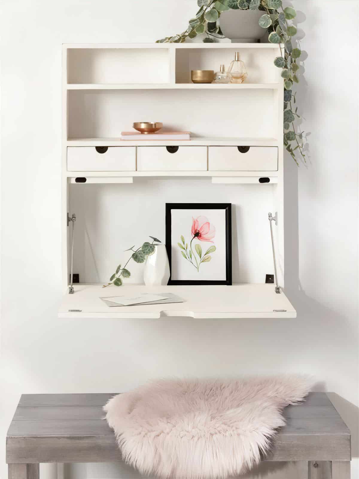 A wall-mounted white desk with small drawers and shelves, decorated with a plant, vases, gold accents, and a framed floral print. Below is a gray bench topped with a light pink faux fur throw.