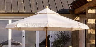 Two cushioned patio chairs with pink pillows are placed on either side of a round wicker side table, all shaded by a large white outdoor umbrella in a sunny garden setting. A stone house and greenery are in the background.