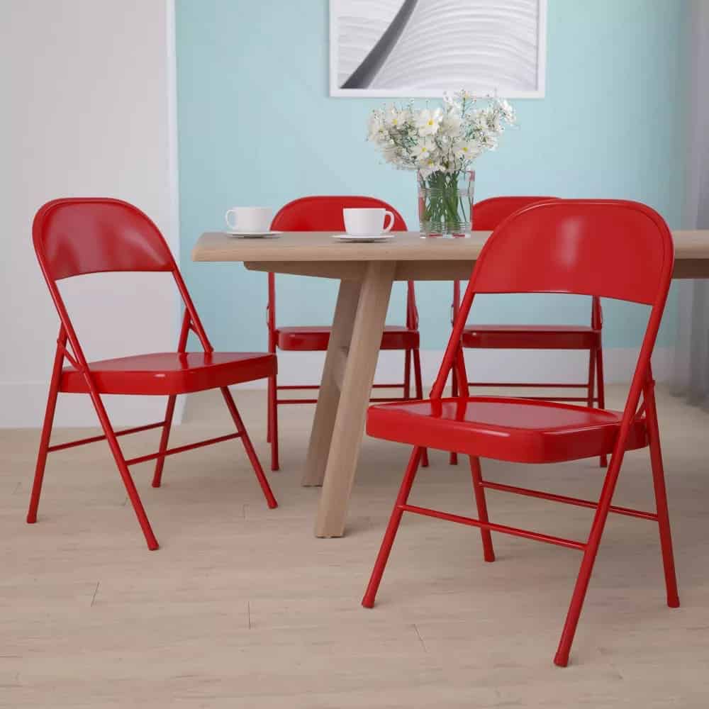 Four bright red metal folding chairs are arranged around a light wooden table with a vase of flowers, two white cups, and a light blue wall in the background.