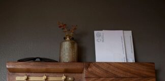 A wooden wall organizer with hooks holding keys, a shelf with sunglasses and a small vase, and a slot containing envelopes, mounted on a gray wall.