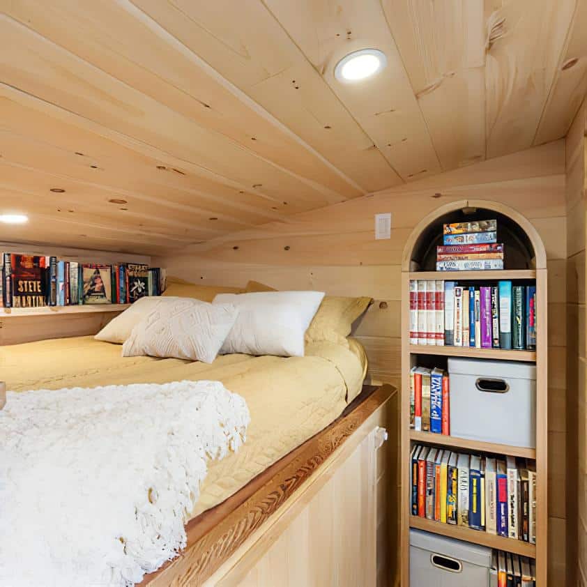 A cozy loft bed with yellow bedding and pillows is nestled under a wooden ceiling, next to shelves filled with books, board games, and storage bins. Recessed lighting brightens the small, warm space.