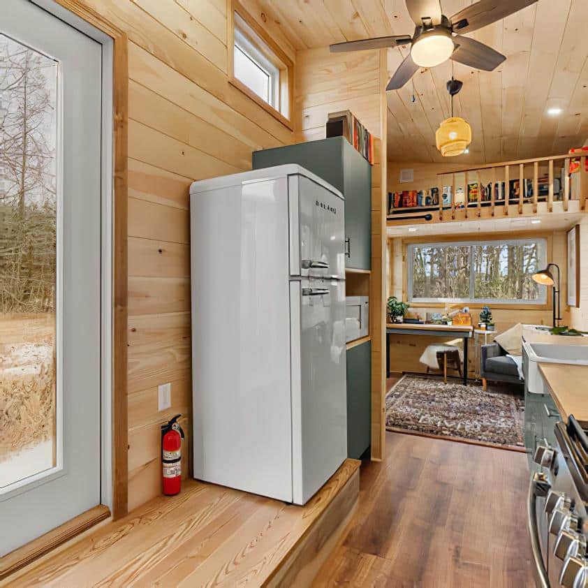 A cozy, modern tiny house interior with wooden walls and floors, a white retro-style fridge, kitchen area, loft with books, ceiling fan, and large windows showing a forest view outside.