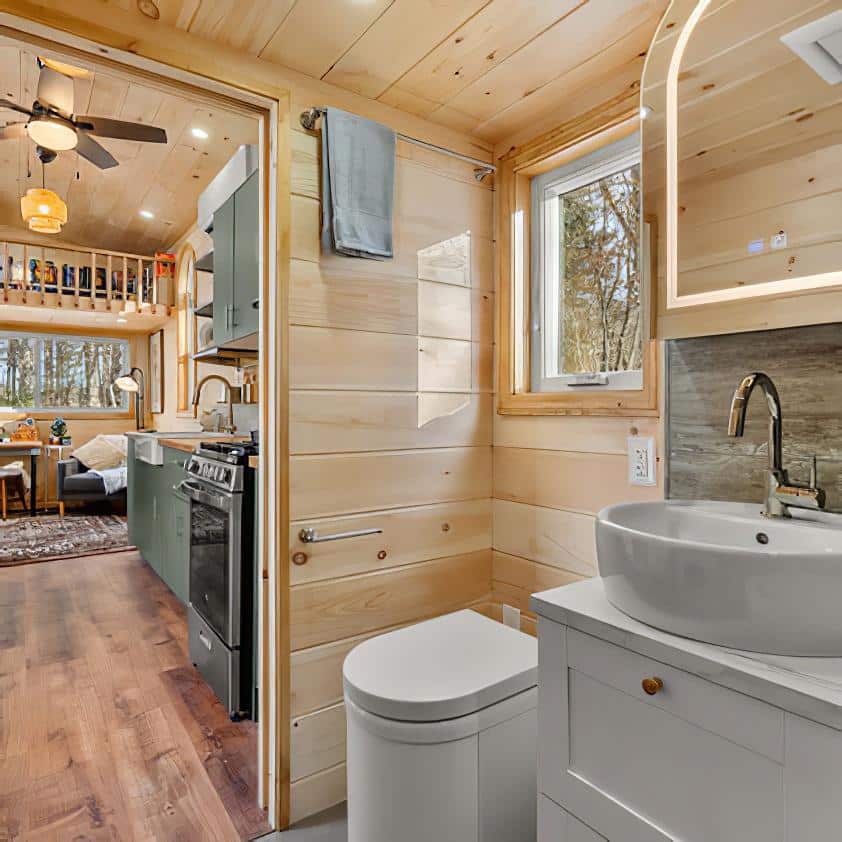 A modern tiny house bathroom with light wood walls, a round sink on a white vanity, a mirror, and a window. The toilet is next to the sink; beyond the bathroom is a small kitchen and cozy living area.
