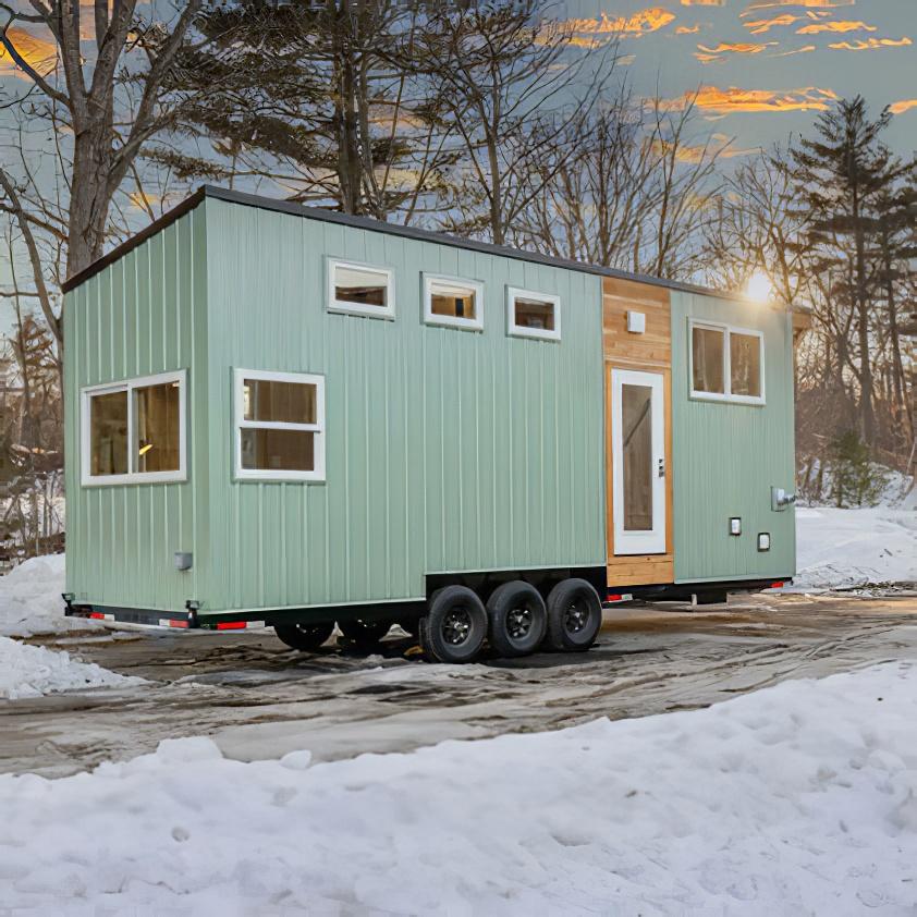 A modern tiny house on wheels with green siding and multiple windows sits on a snowy driveway, surrounded by bare trees under a partly cloudy sky at sunset.