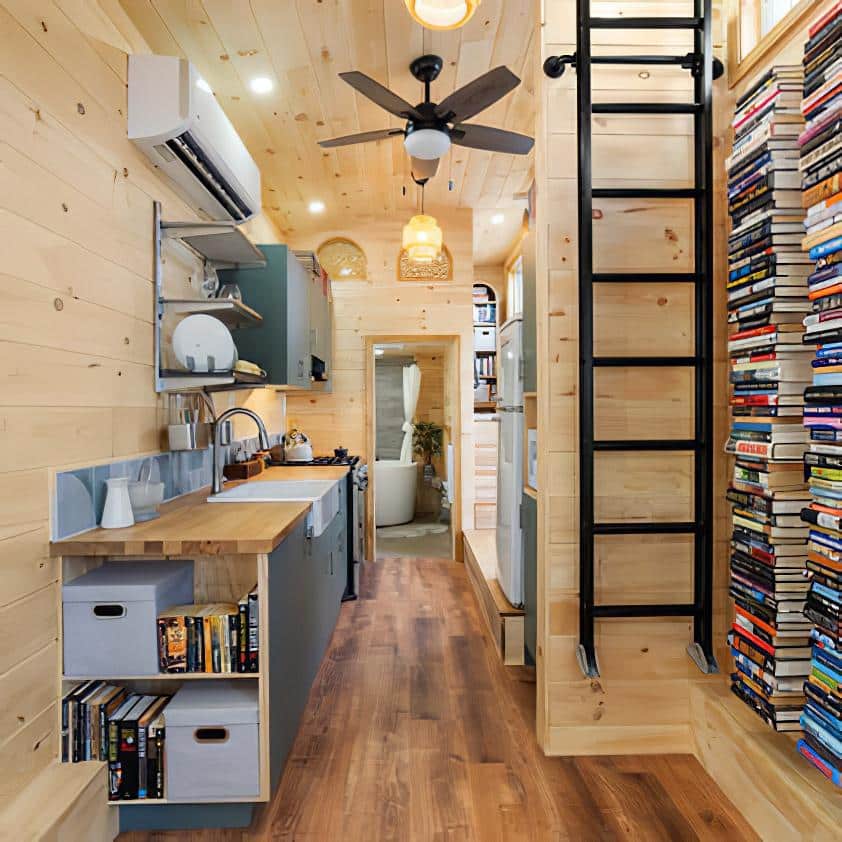 A cozy, modern tiny house interior with wooden walls and floors. A kitchen with shelves and appliances is on the left, a ladder leads to a loft, and a wall-mounted bookshelf filled with books is on the right.