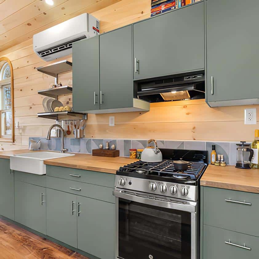 A modern kitchen with sage green cabinets, wood countertops, a stainless steel gas stove, a farmhouse sink, open shelves, and light wood panel walls. A teapot sits on the stove and kitchen items are neatly arranged.