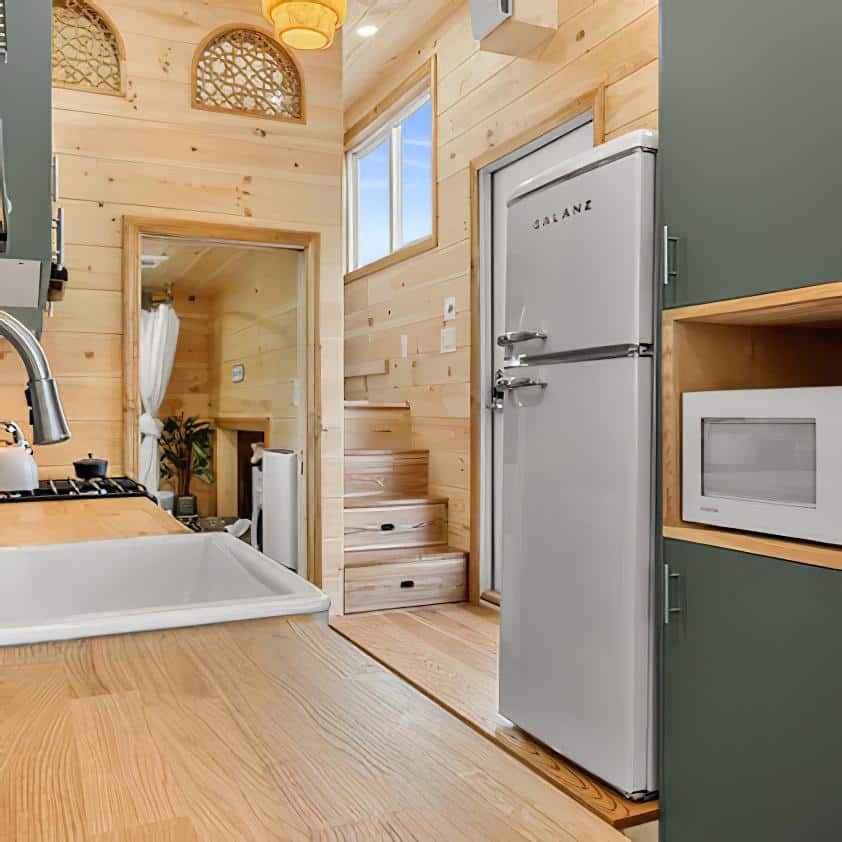 A compact, modern tiny house kitchen with green cabinets, a white retro-style refrigerator, a microwave, a farmhouse sink, and wood paneled walls leading to a staircase and a bright loft area.