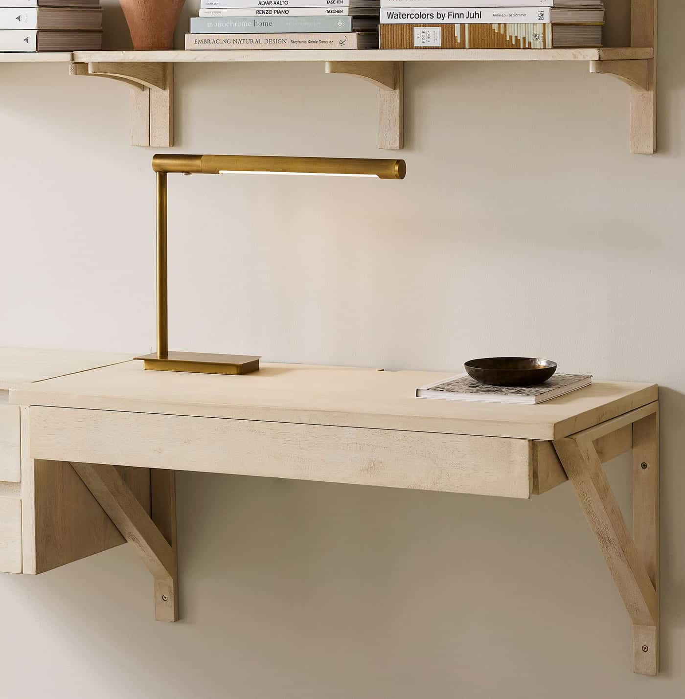 A minimalist wooden wall-mounted desk with an open drawer, a gold desk lamp, a dark bowl, and a closed notebook on top. Books are stacked on wooden shelves above the desk against a light-colored wall.