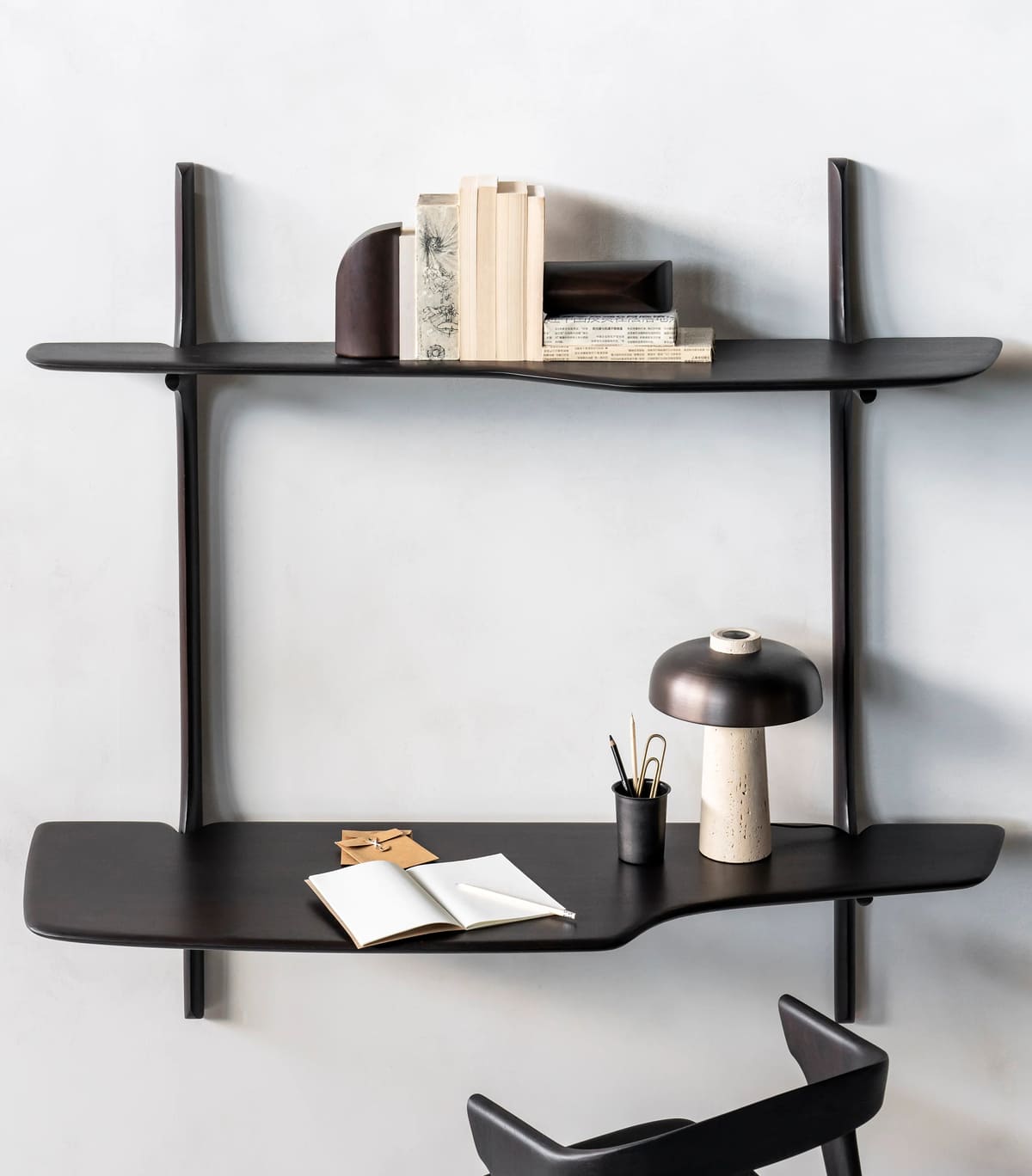 Two modern, dark wood wall-mounted shelves hold books, decorative objects, a table lamp, an open notebook, and writing utensils. A black chair is partially visible in front of the lower shelf.