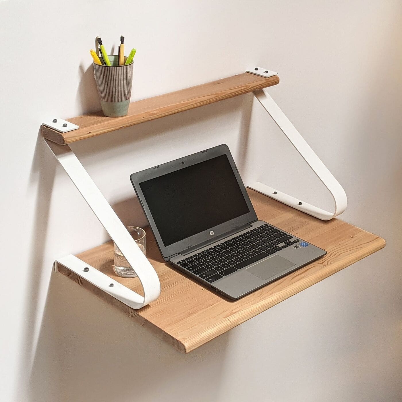 A minimalist wall-mounted wooden desk with white metal brackets, holding a laptop, a glass of water, and a cup with pens and pencils on an upper shelf, against a plain white wall.