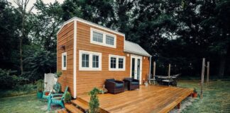 A modern tiny house with wood siding sits on a spacious wooden deck, featuring outdoor seating, potted plants, and surrounded by grass and trees under an overcast sky.