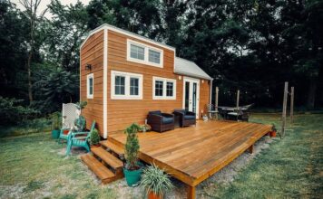 A modern tiny house with wood siding sits on a spacious wooden deck, featuring outdoor seating, potted plants, and surrounded by grass and trees under an overcast sky.