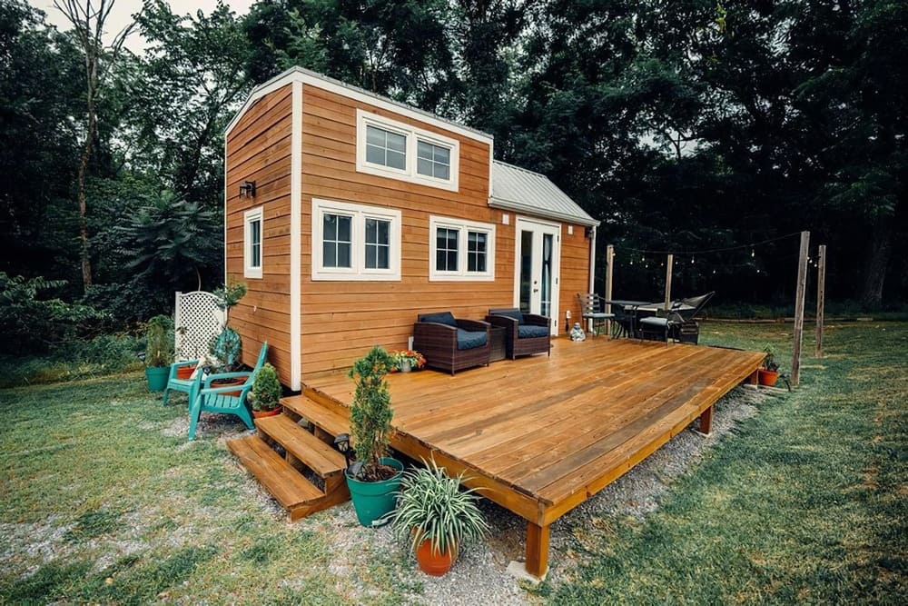 A modern tiny house with wood siding sits on a spacious wooden deck, featuring outdoor seating, potted plants, and surrounded by grass and trees under an overcast sky.