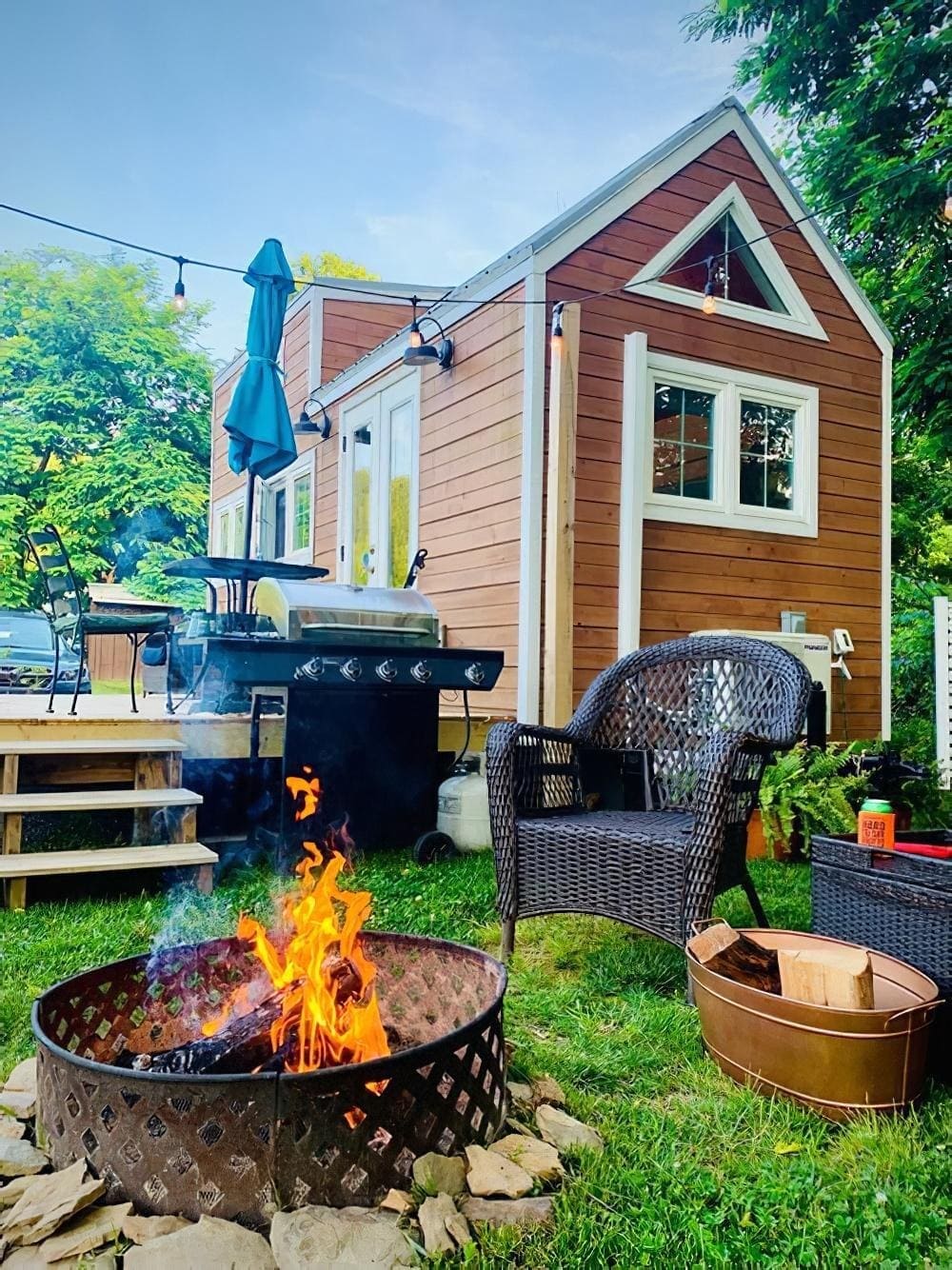A cozy backyard scene with a small wooden tiny house, a fire pit with a burning fire, wicker chairs, a grill, and a patio dining set surrounded by green grass and trees.
