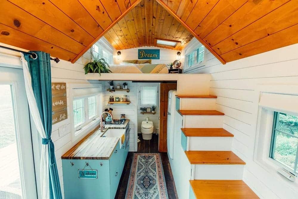 Cozy tiny house interior with wood-paneled ceiling, white walls, teal cabinets, a small kitchen, steps leading to a loft, narrow rug, and a compact bathroom at the back. Bright natural light fills the space.
