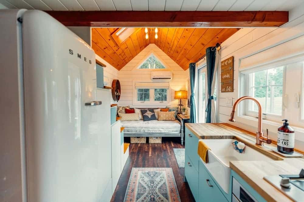 A cozy, well-lit tiny house interior with wood accents, a modern kitchen including a farmhouse sink, a retro-style refrigerator, and a sitting area with a cushioned bench and decorative pillows by large windows.
