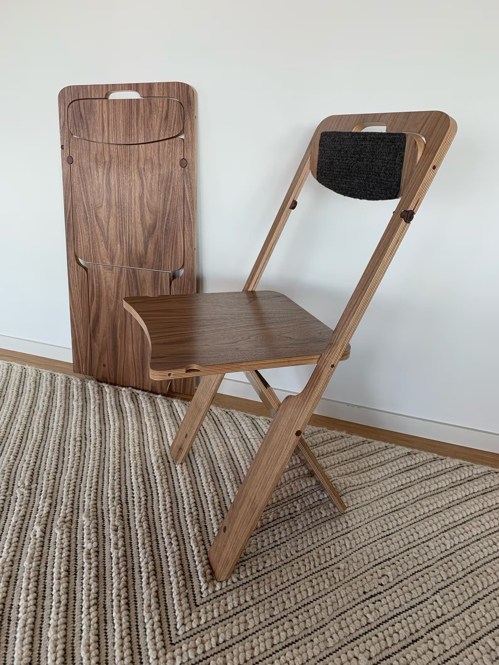 A wooden foldable chair with a black cushion on the backrest sits open on a textured rug, while another identical chair is folded and standing upright against a white wall.