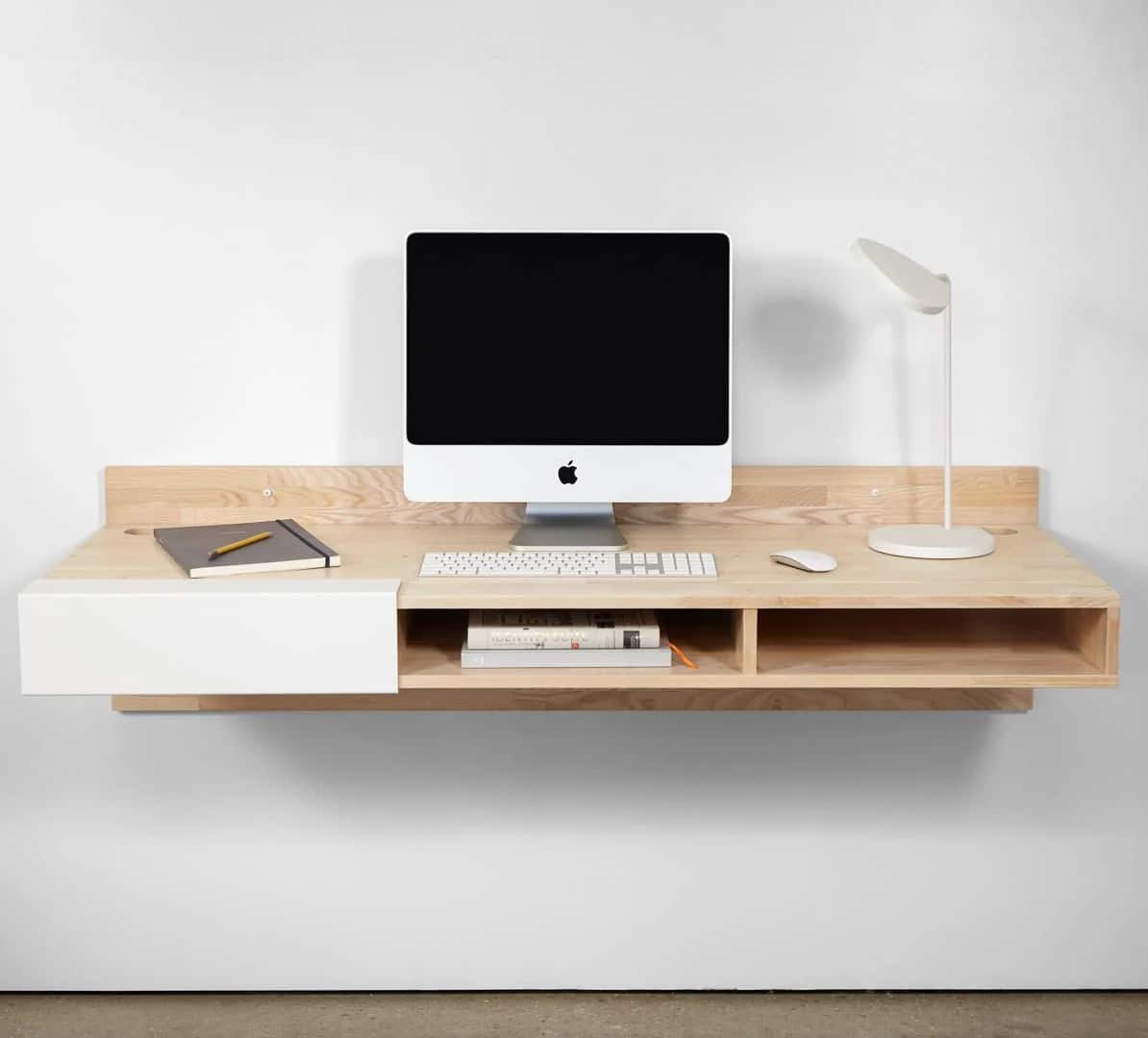 A minimalist floating wooden desk with two open shelves and a white drawer, featuring an iMac, keyboard, notebook, pencil, closed book, and a modern white desk lamp against a plain white wall.