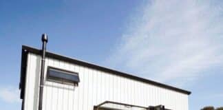 A modern white tiny house with black trim sits on a grassy lawn. It has a small porch with steps, a potted plant, and large windows. The sky is blue with wispy clouds.