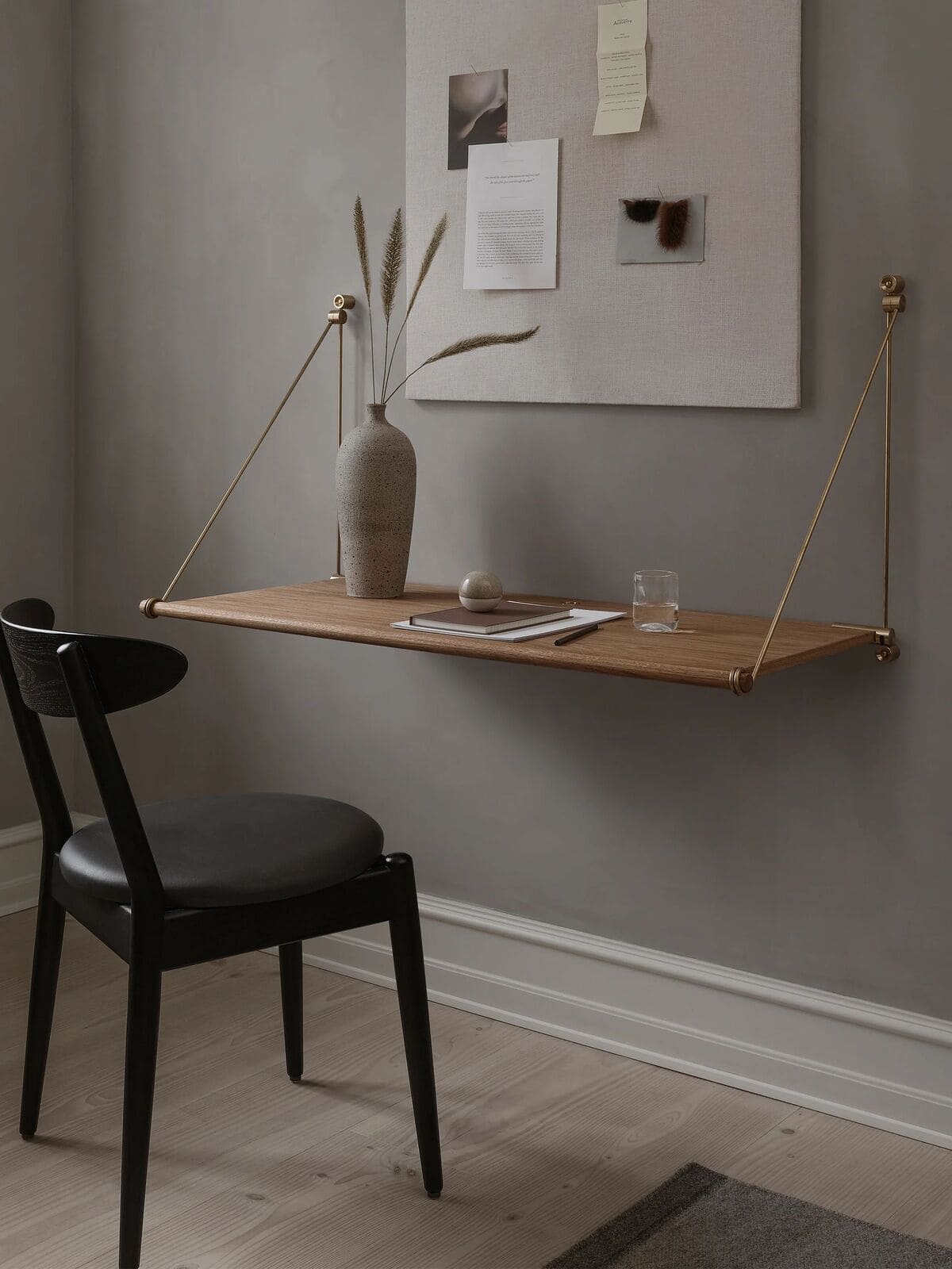 A minimalist workspace with a black chair, a floating wooden desk supported by brass rods, a ceramic vase with dried grass, a glass of water, and papers, set against a gray wall with a pinboard.