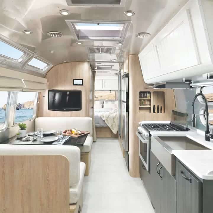 Interior of a modern Airstream RV featuring a dining area, kitchen, and bed; Pottery-Barn-inspired light wood cabinetry, stainless steel appliances, large windows with an ocean view, and bright, clean decor.